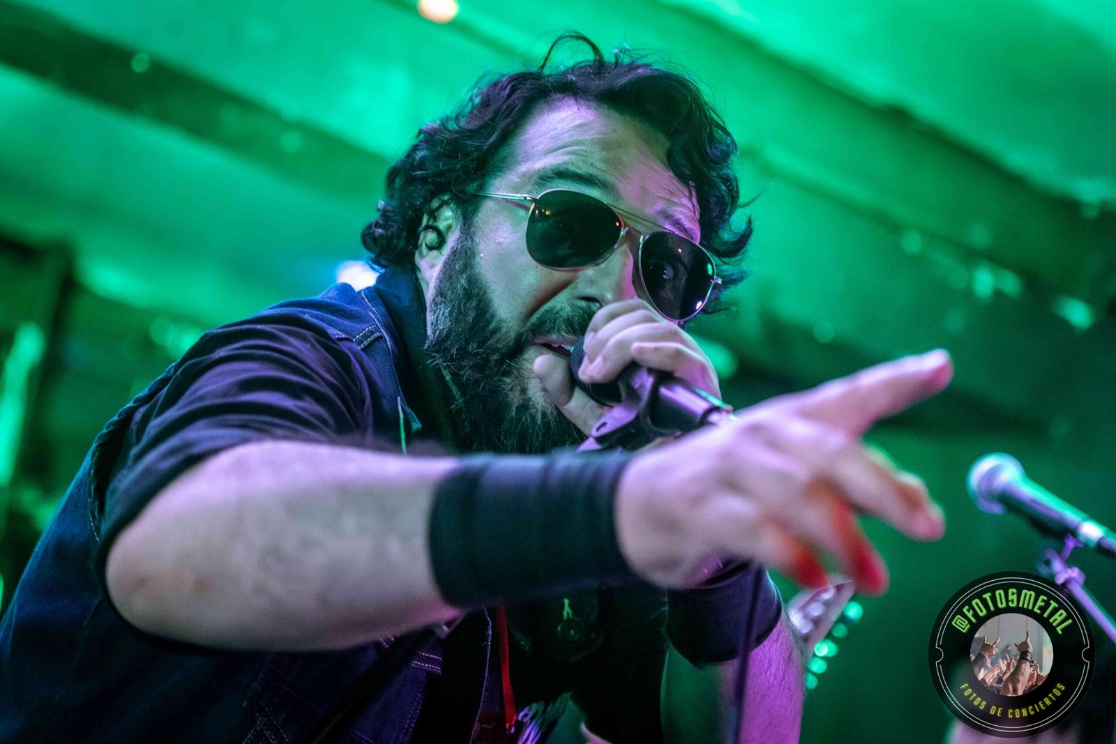 Cantante con gafas y barba en escenario iluminado de verde apuntando y sosteniendo micrófono.