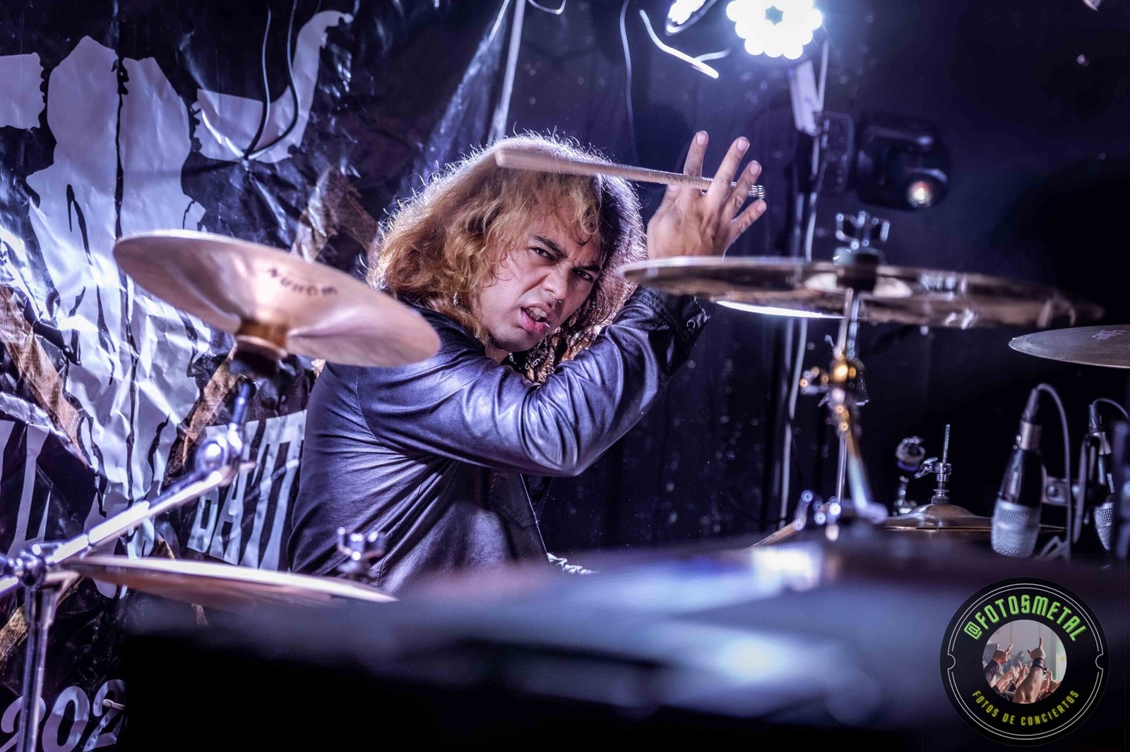 Baterista con chaqueta de cuero tocando batería en escenario iluminado durante concierto.