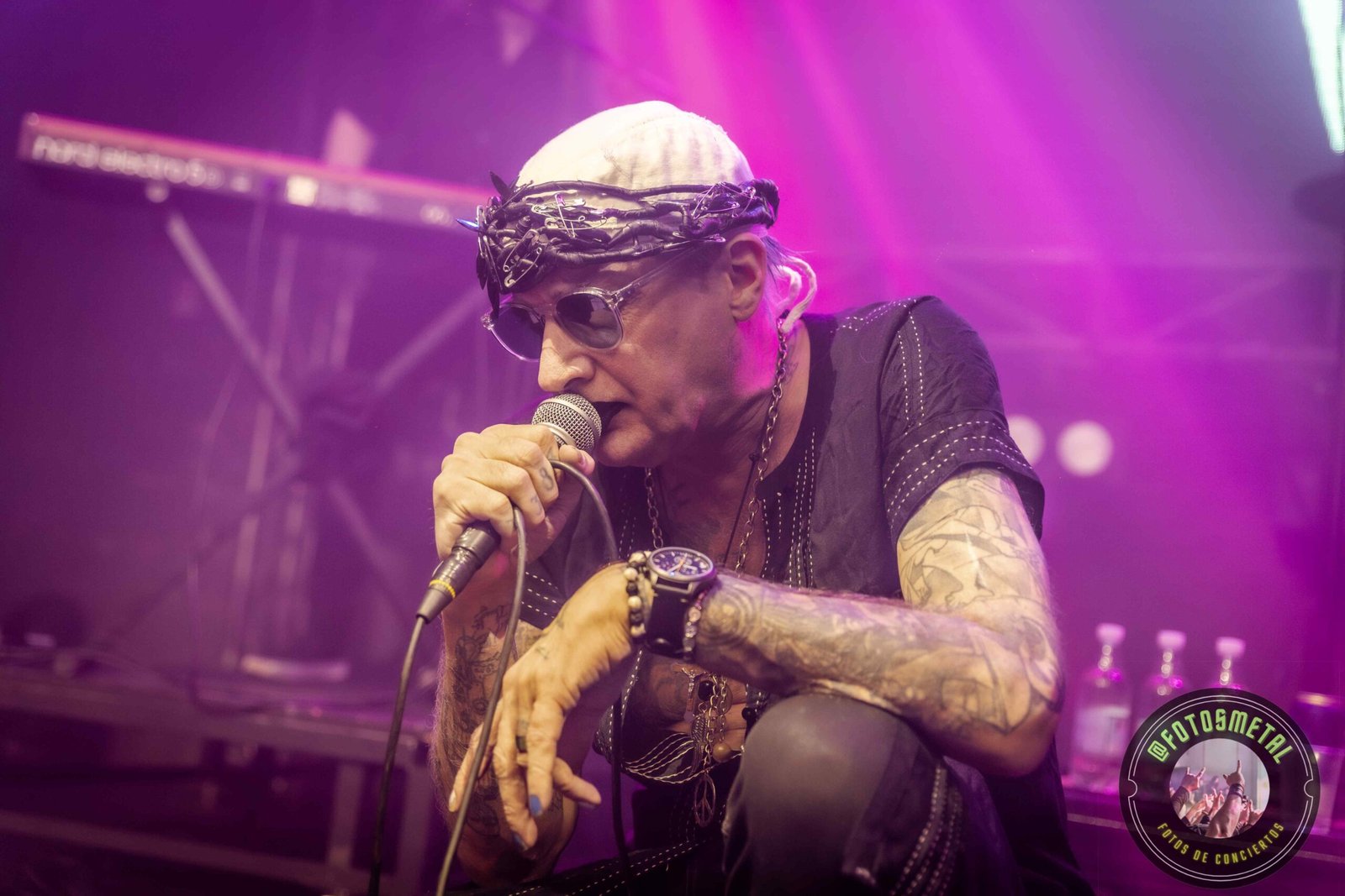Cantante con gafas y barba en escenario iluminado de verde apuntando y sosteniendo micrófono.