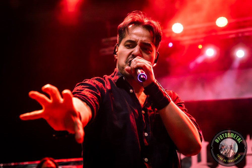 Cantante con micrófono en mano durante actuación, mano extendida hacia cámara, luces rojas y humo.