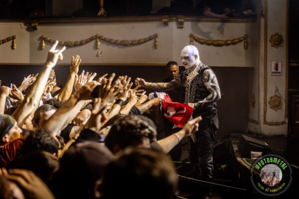 Músico maquillado en blanco y vestimenta oscura entrega prenda roja y blanca a la multitud desde el borde del escenario.