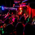 Concierto de metal en bar: banda en escenario con guitarras, bajo y batería, público grabando y viendo. Iluminación roja, verde y azul. Amplificadores Marshall y pantallas detrás. Ambience oscuro y