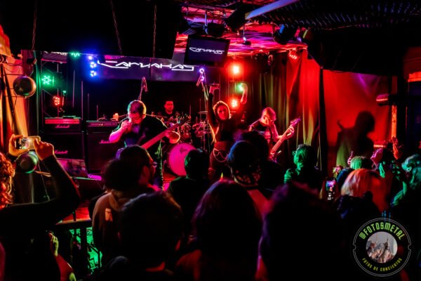 Concierto de metal en bar: banda en escenario con guitarras, bajo y batería, público grabando y viendo. Iluminación roja, verde y azul. Amplificadores Marshall y pantallas detrás. Ambience oscuro y